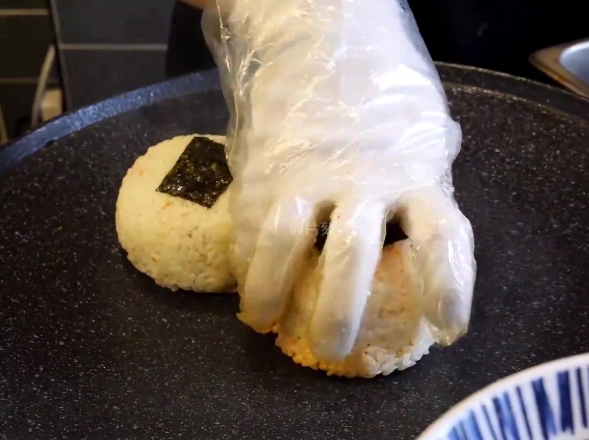 A close-up of a gloved hand pressing seasoned rice balls onto a hot, black griddle to sear them.