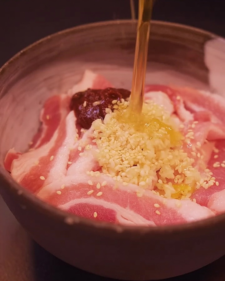 Pork belly slices in a bowl being seasoned with gochujang, minced garlic, sesame seeds, and sesame oil.