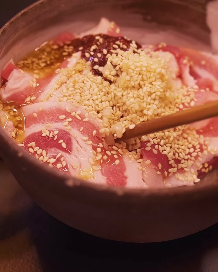 Close-up of chopsticks mixing marinated pork belly slices in a bowl.