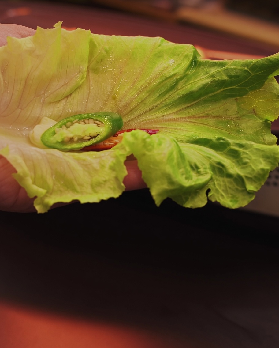 A hand holding a fresh lettuce leaf with a slice of grilled pork belly and chili ready for assembly.
