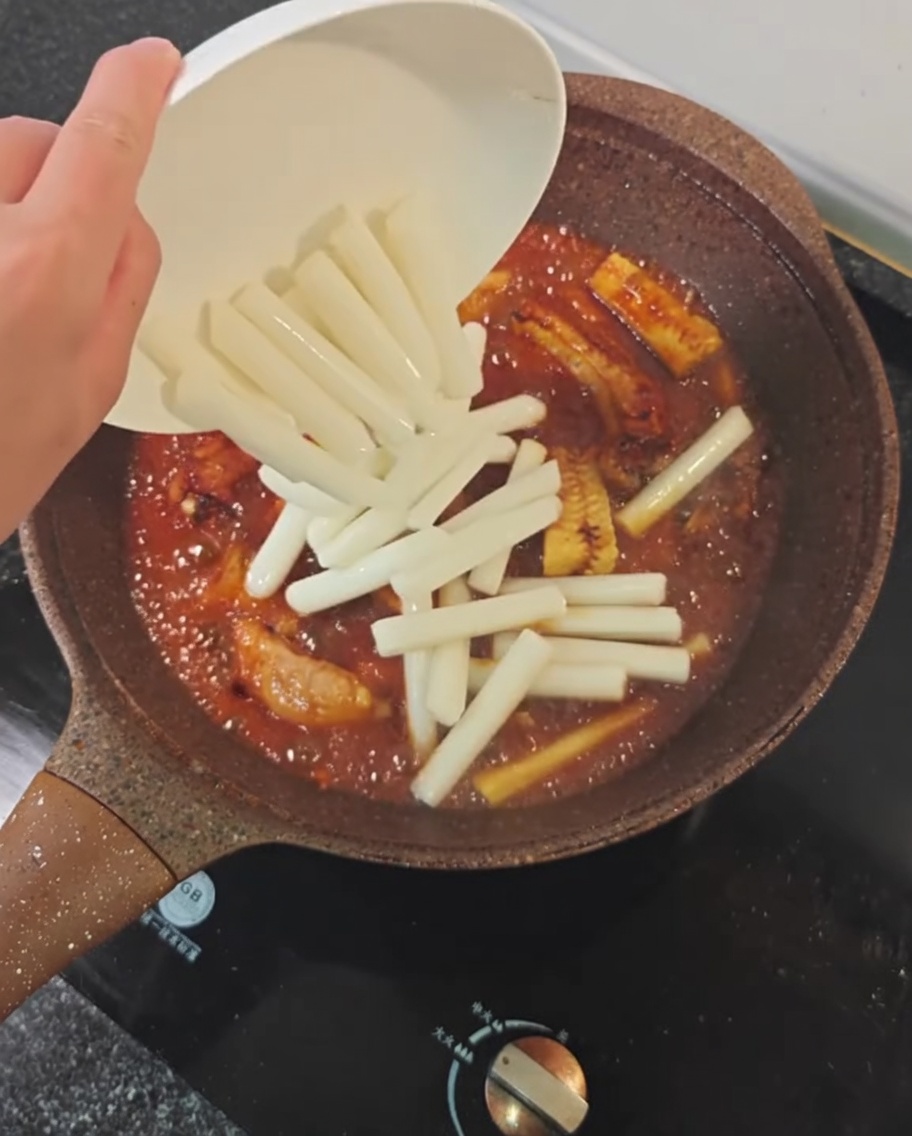 Rice cakes being dropped into a pan containing pork ribs, corn, and spicy braising sauce.