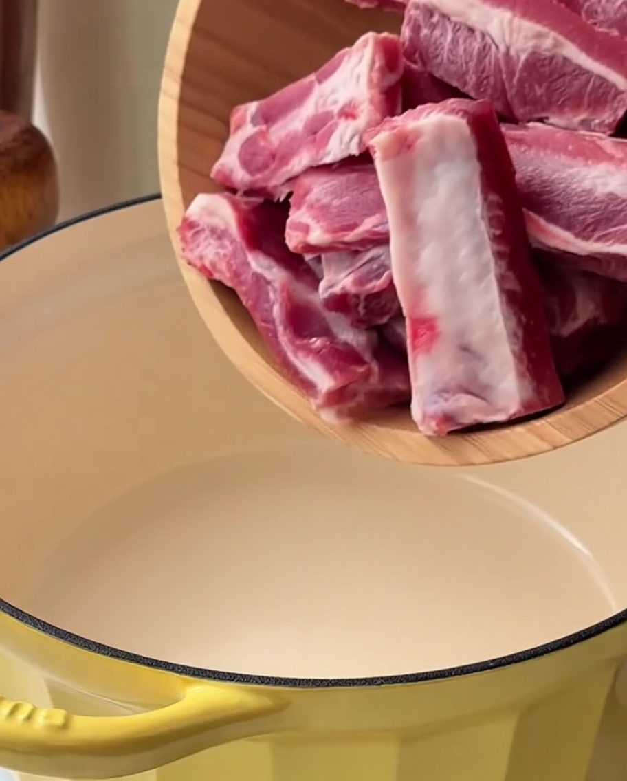 Pork rib pieces in a yellow pot submerged in water with green onion and ginger, ready to be blanched.