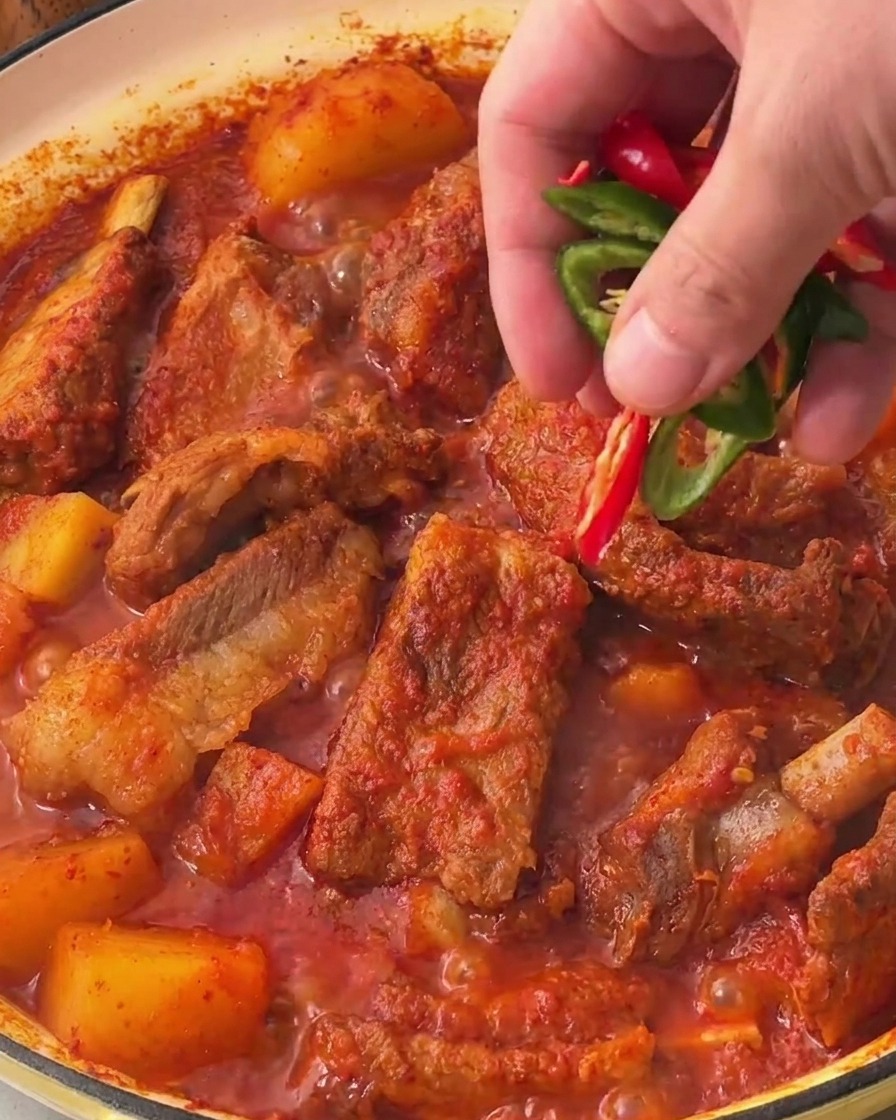 A hand scattering fresh, bright green and red chili pepper slices over the surface of the finished Korean spicy stewed pork ribs.