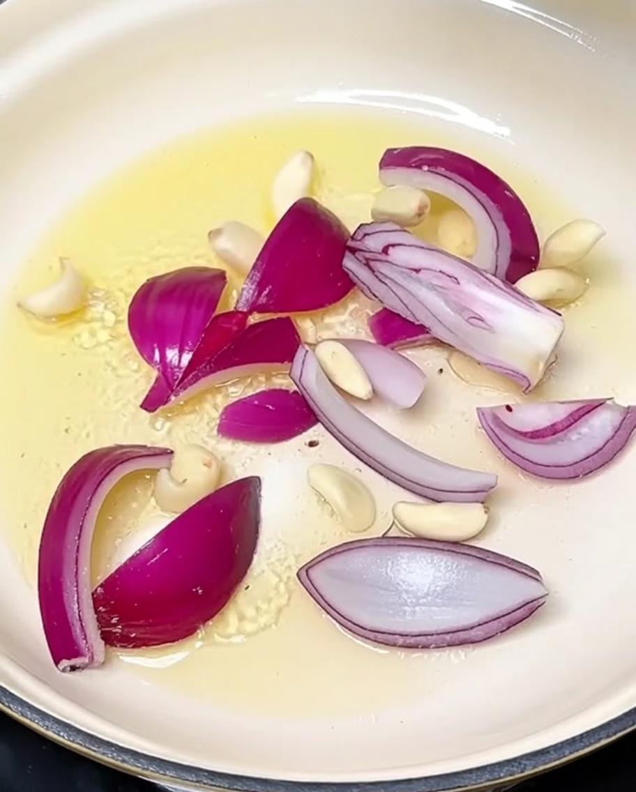 Sliced onions and garlic cloves being sautéed in hot oil in a cooking pot.