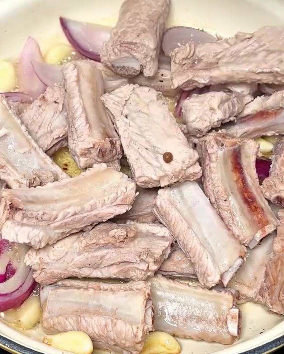 Pork ribs being sautéed in a pot alongside garlic cloves and sliced red onions.