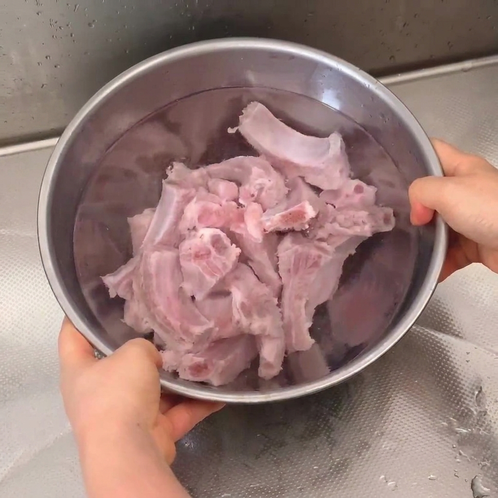 Fresh pork ribs soaking and being rinsed in a stainless steel bowl under a kitchen faucet.