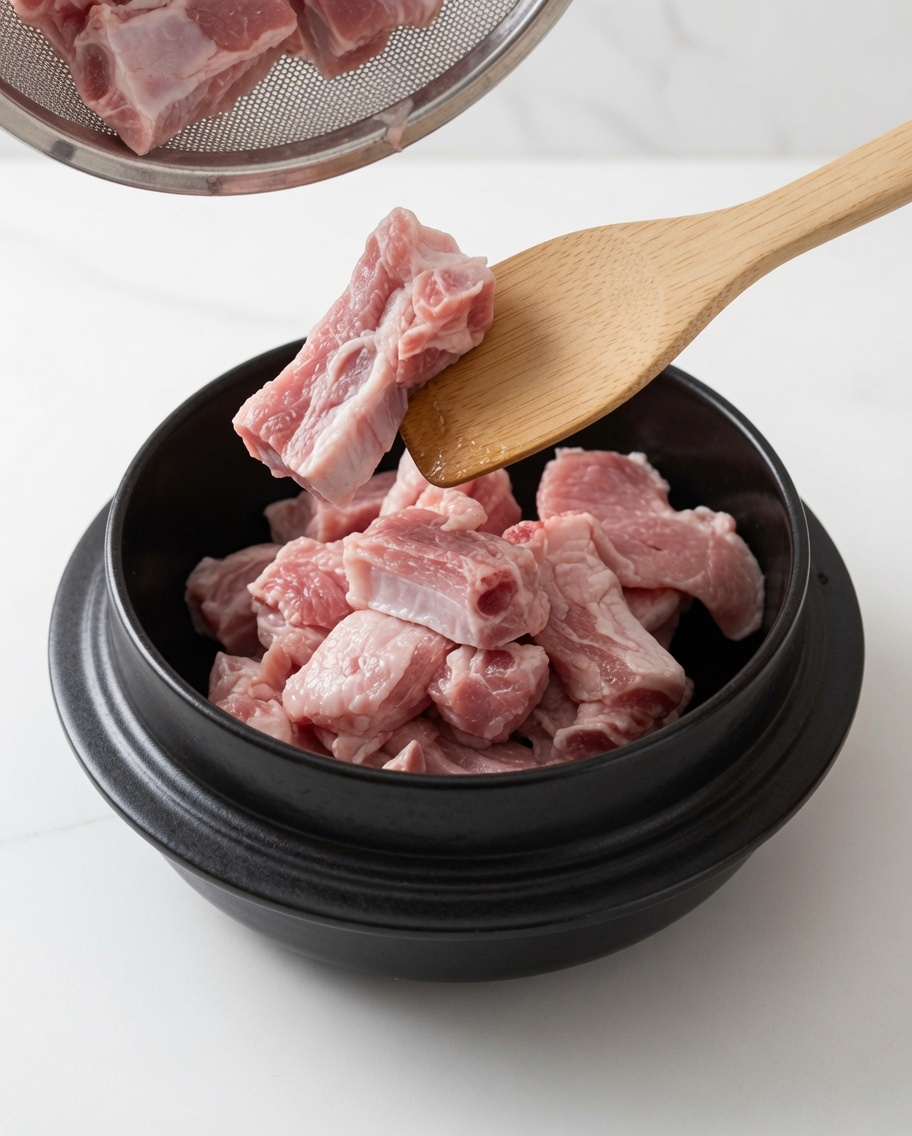 Pork ribs being stir-fried in a pot with oil and white sugar using a wooden spoon.
