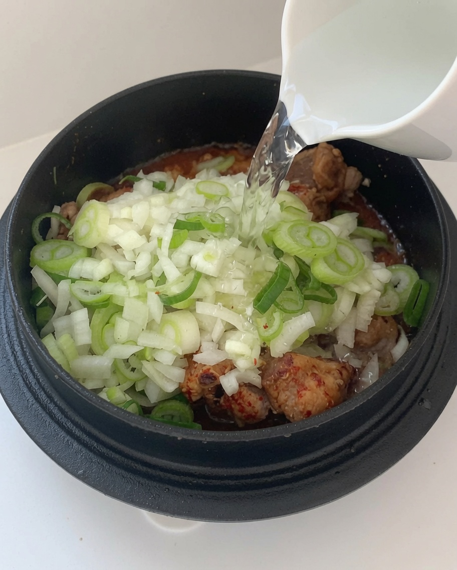 Water being poured from a white pitcher into a pot filled with seasoned pork ribs and aromatics.