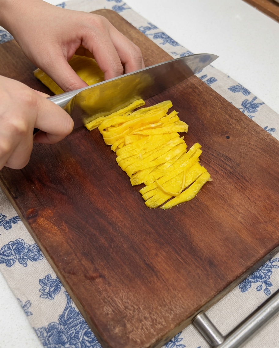 A close-up view of a hand using a kitchen knife to slice a yellow, cooked thin egg crepe into neat strips on a wooden cutting board.