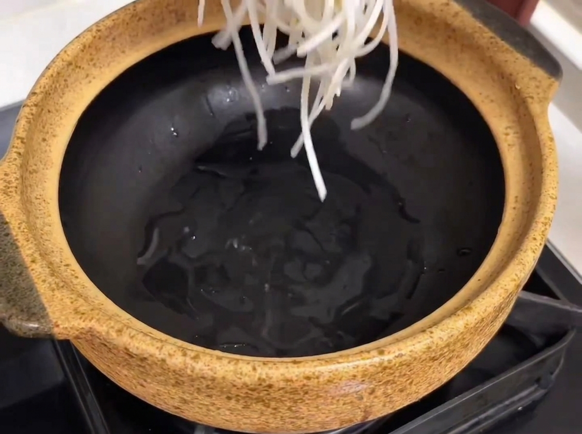 A ceramic serving pot being prepared with glass noodles, tender beef ribs, and chunks of radish placed inside.