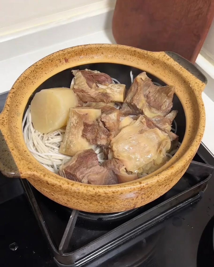A clear, rich beef bone broth being poured from a rice cooker into a ceramic pot filled with beef ribs, radish, and noodles.