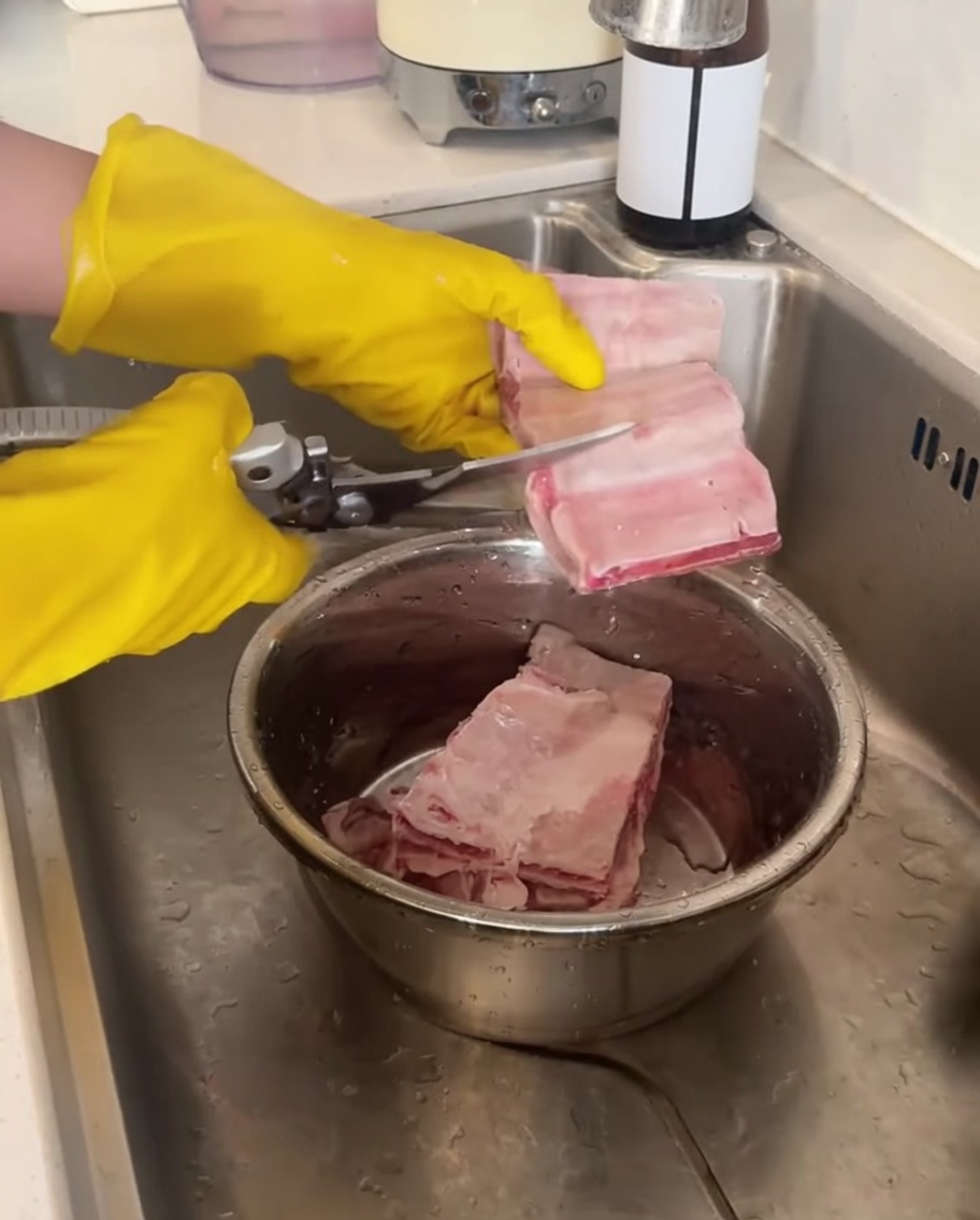 A pair of yellow-gloved hands using kitchen shears to cut raw beef ribs over a metal bowl.