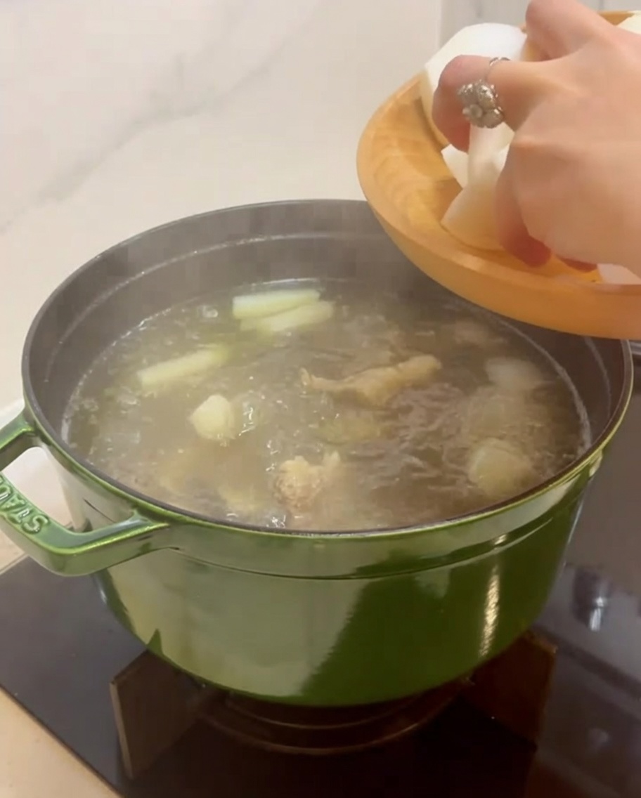 Hand adding large white chunks of peeled Korean radish into a simmering pot of beef soup.