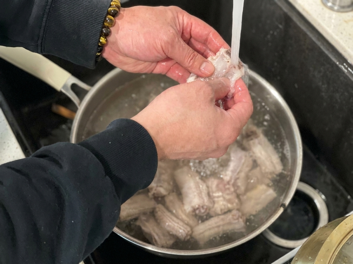 Blanched pork ribs being rinsed under a kitchen faucet with running water.