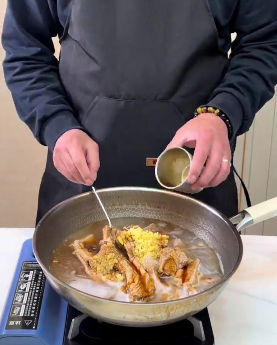 Freshly prepared pear juice being poured from a small metal cup into a cooking pot with ribs and seasonings.