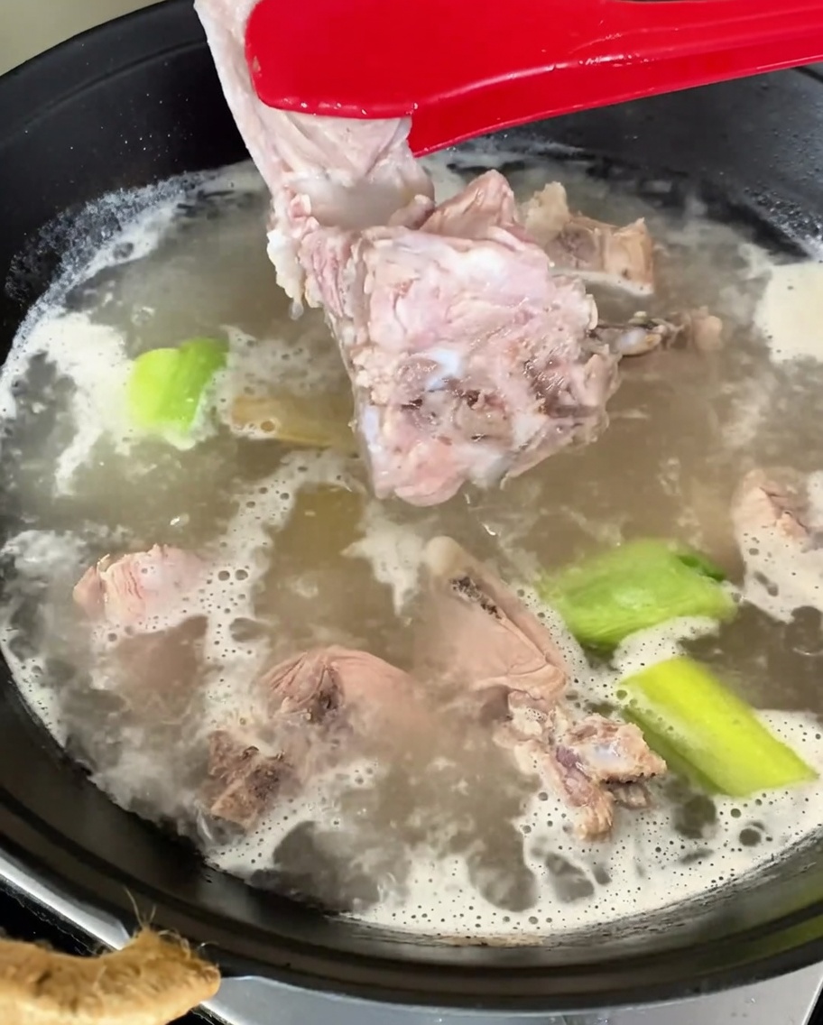 Blanched pork spine pieces being lifted from boiling water with red tongs.