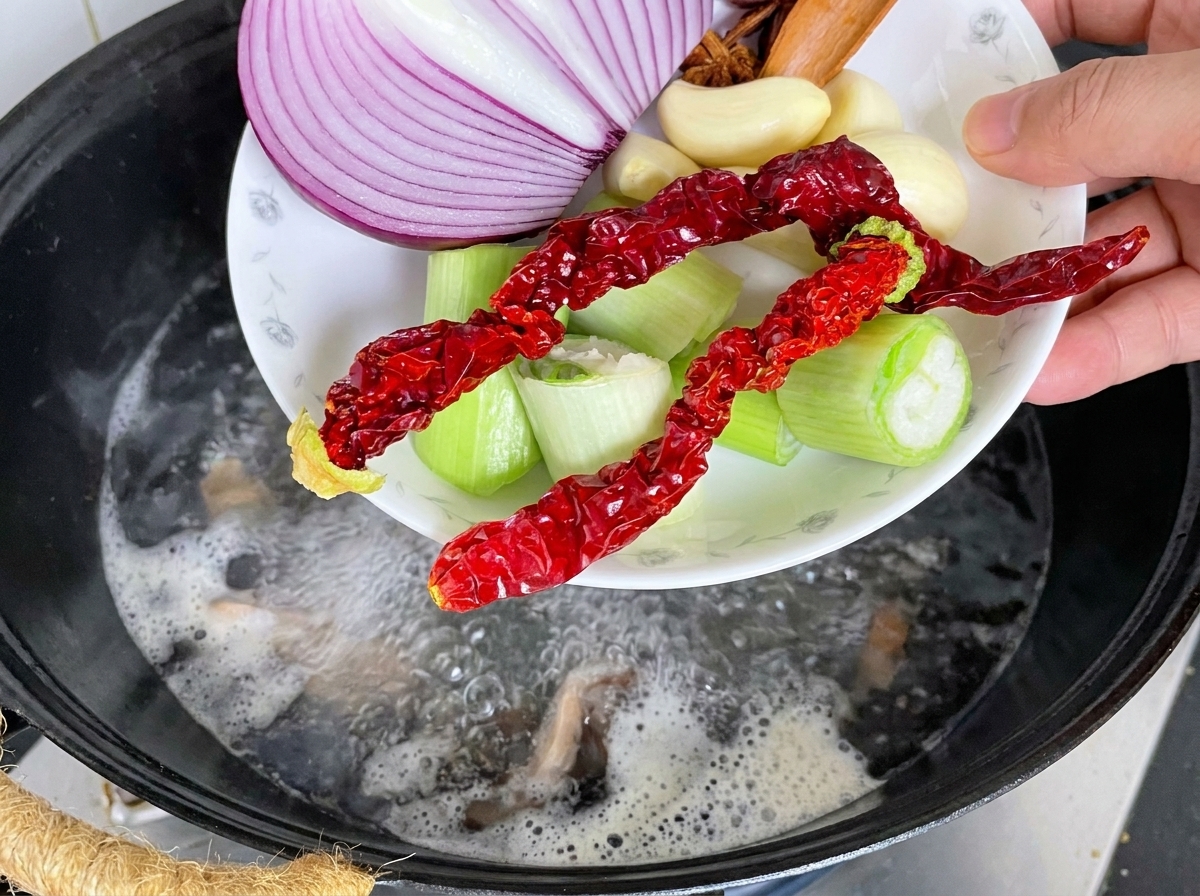 Pork spine in a wok with aromatics and boiling water being poured in.