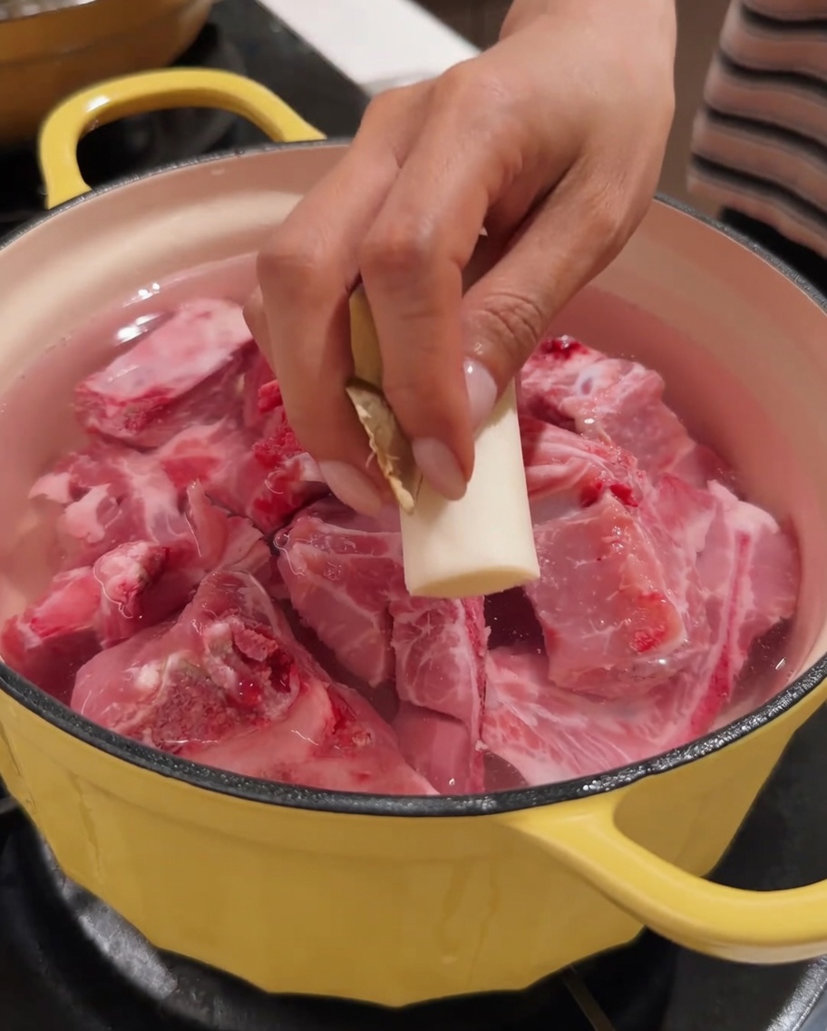 A hand placing a piece of ginger into a pot containing pork spine and water.