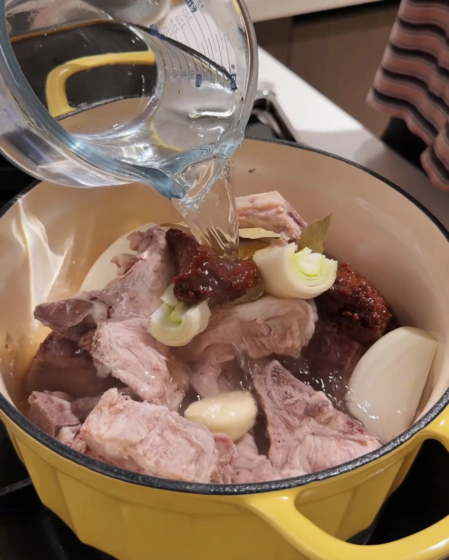 Hot water being poured from a measuring cup into the pot with pork and seasonings.