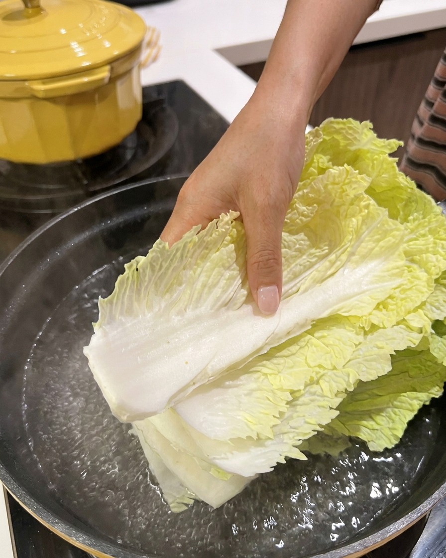 Fresh napa cabbage leaves being lowered into a pot of boiling water on the stove.