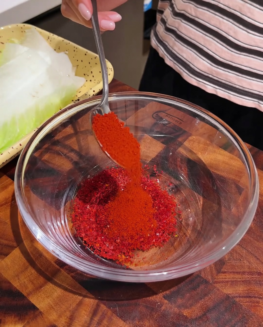 Red chili powder being spooned into a clear glass bowl containing other seasoning spices.