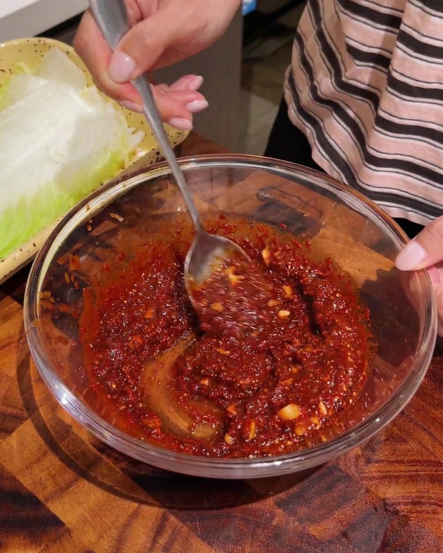 Torn strips of blanched napa cabbage being mixed with a red seasoning sauce in a glass bowl.