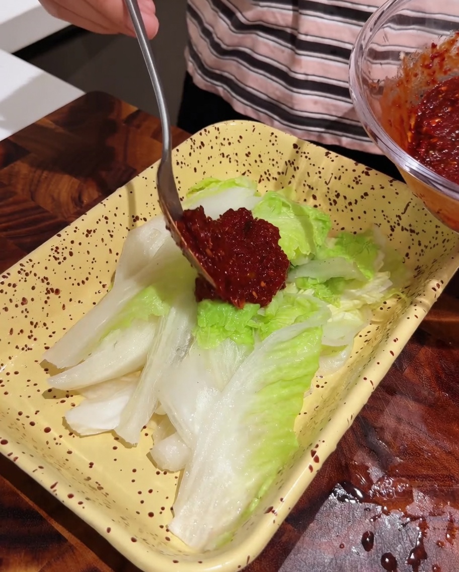 Spoon applying red seasoning paste onto a tray of fresh cabbage leaves.