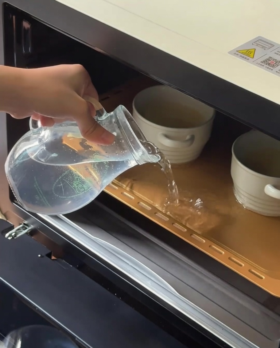 Warm water being poured from a glass pitcher into a baking tray containing ceramic ramekins.
