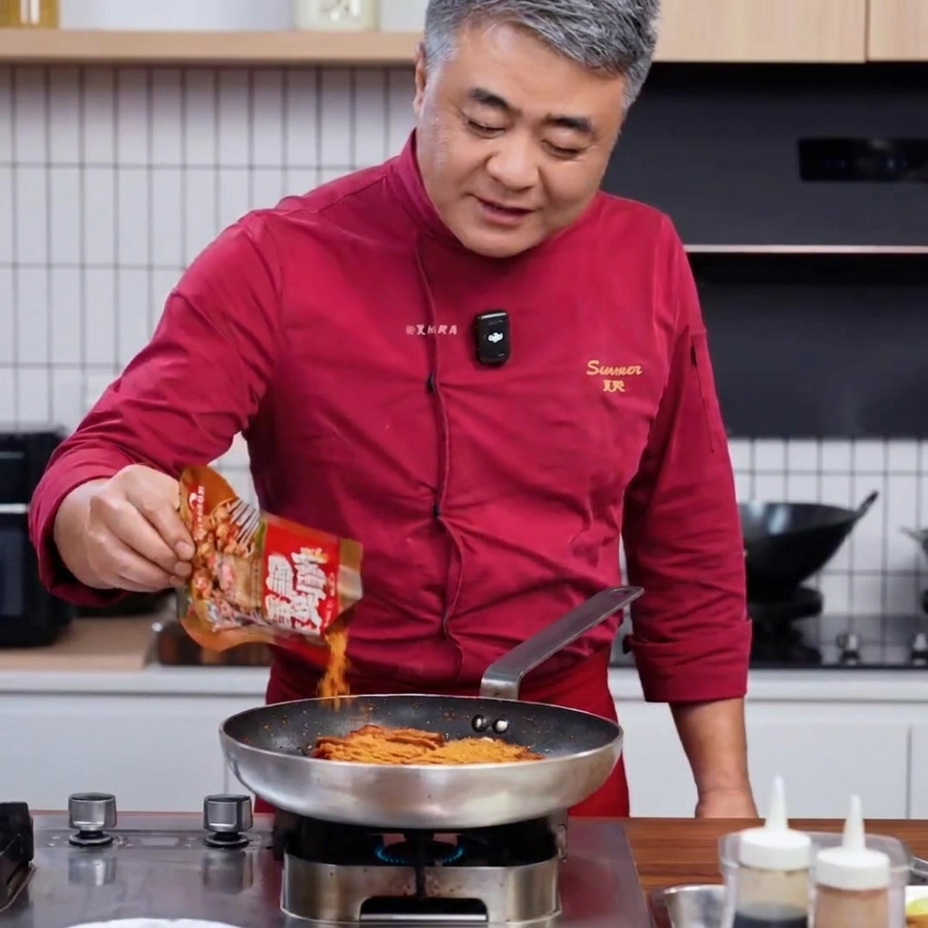 Chef in a red coat pouring barbecue spice mix from a pouch into a pan of seared pork.