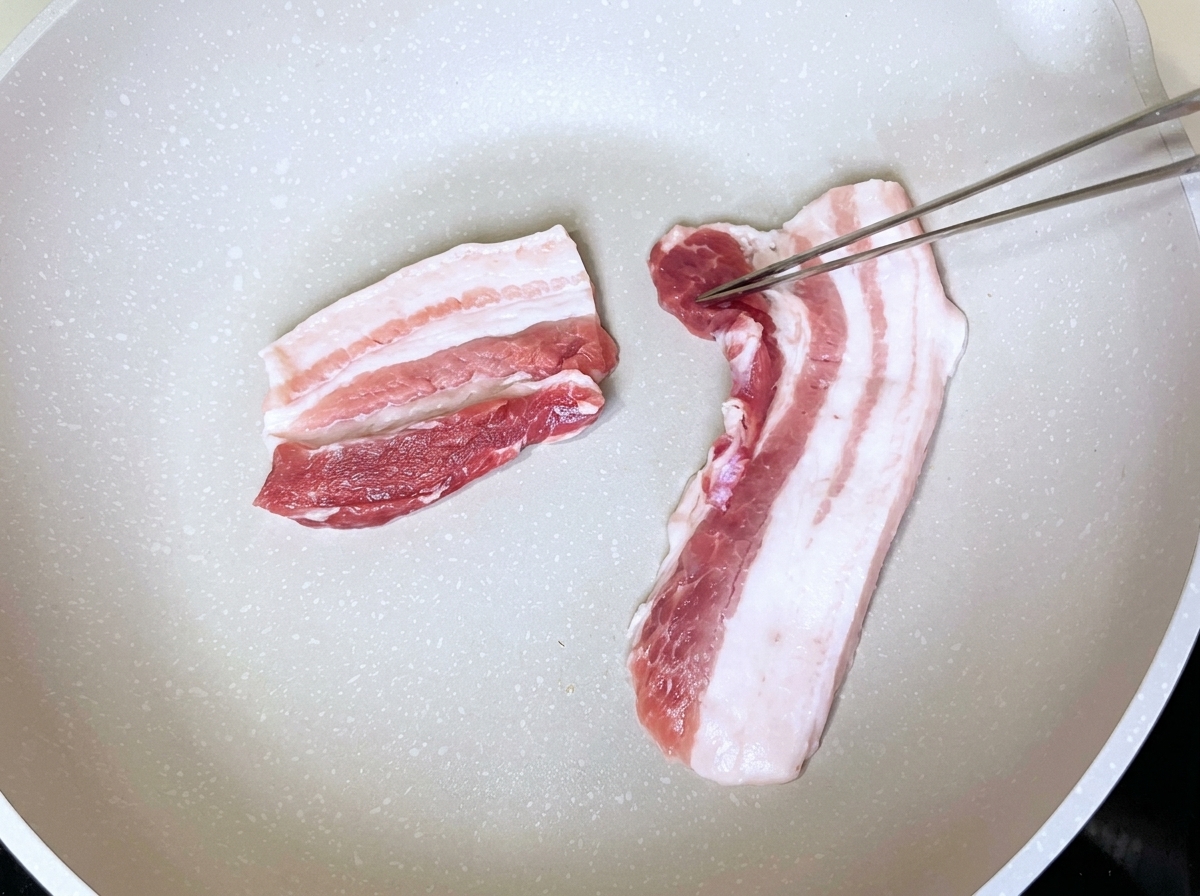 Pork belly slices placed neatly in a dry white pan being handled with metal tongs.