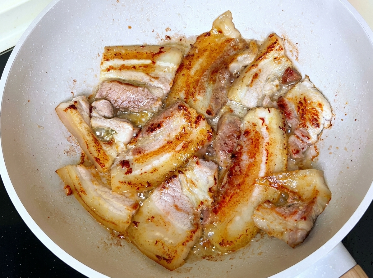 Slices of pork belly frying and browning in a pan with rendered fat.