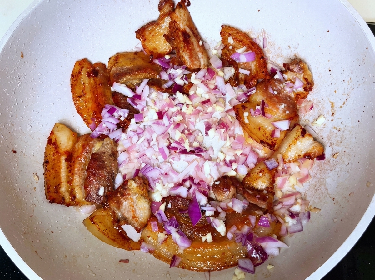 Diced red onions and minced garlic scattered over browned pork pieces in a white pan.