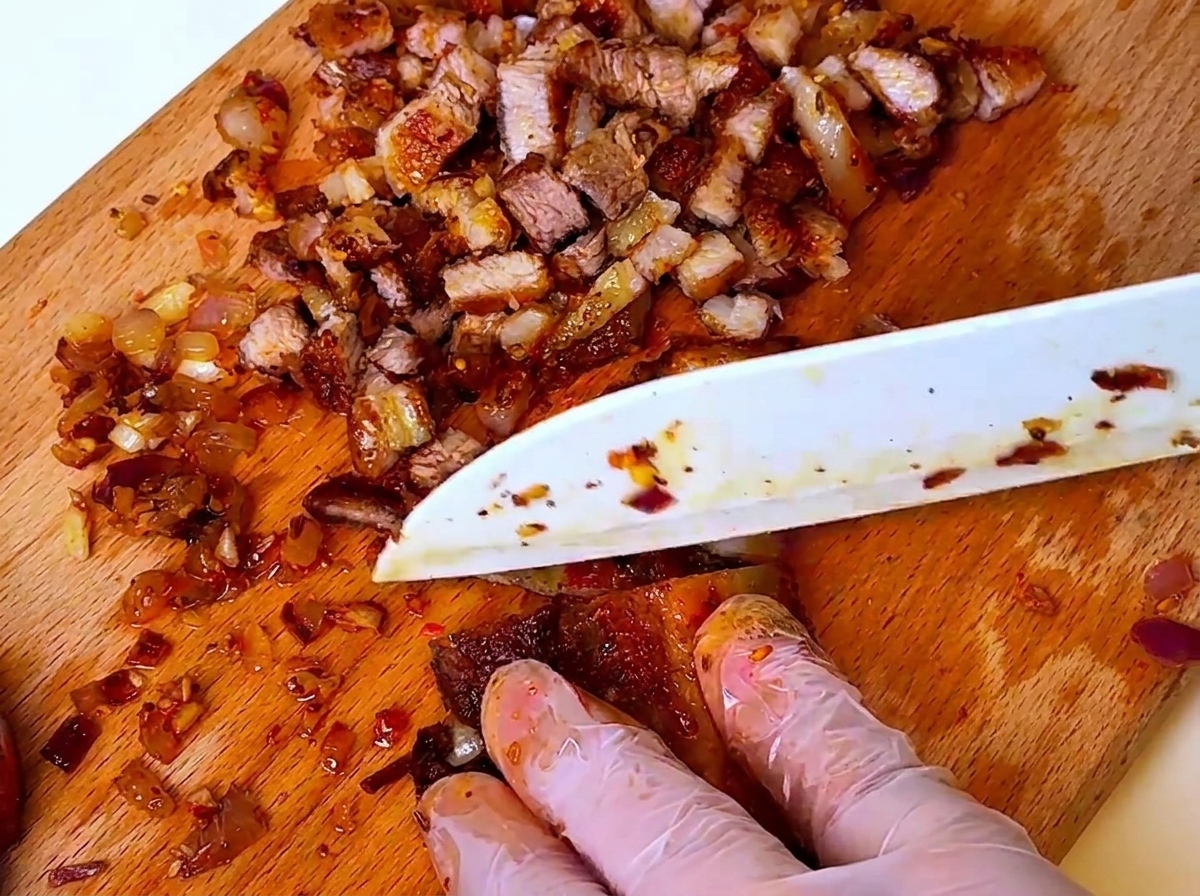 A knife chopping cooked barbecue pork and onions on a wooden cutting board, with hands wearing protective gloves.