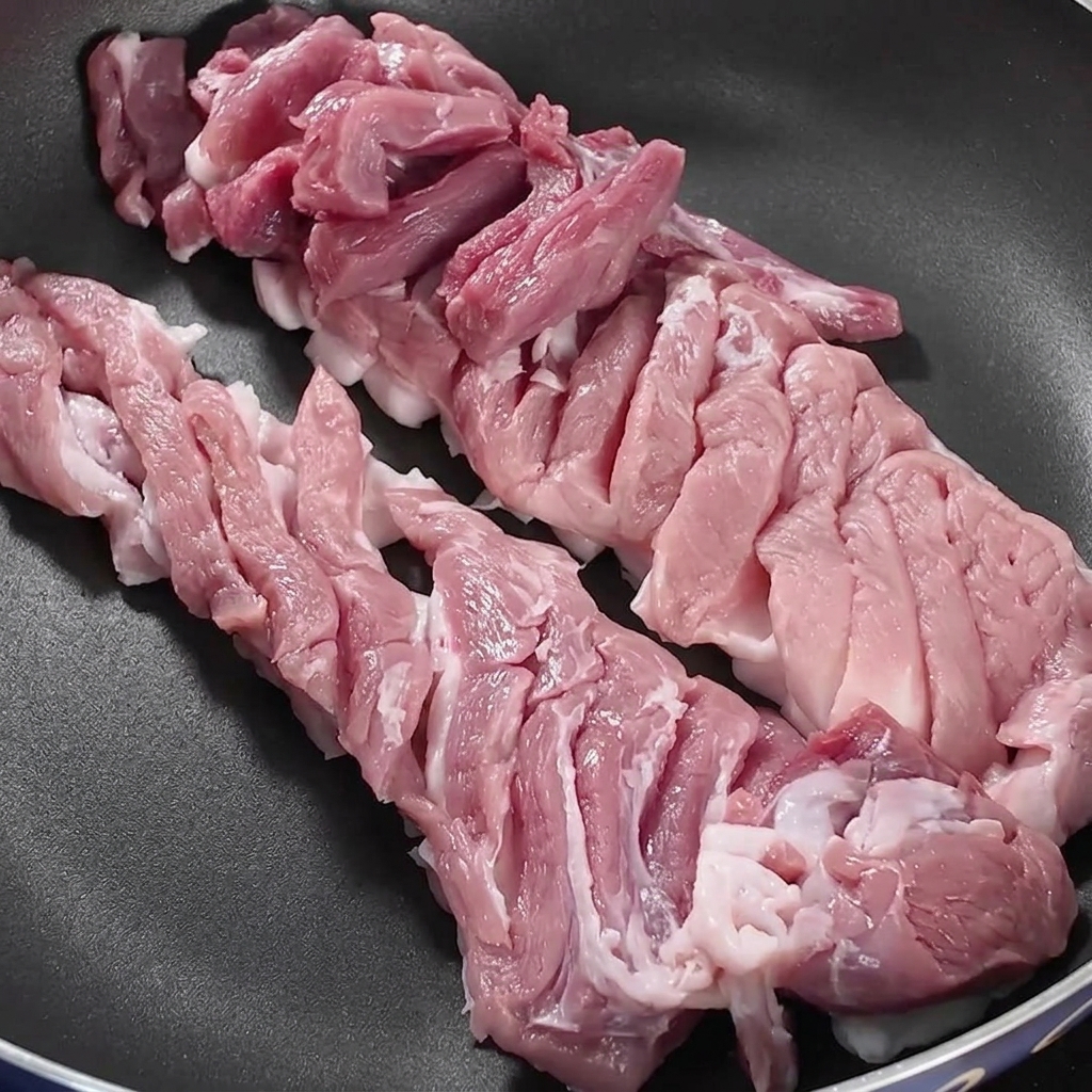 Slices of pork belly arranged neatly inside a dark frying pan over a stovetop burner.