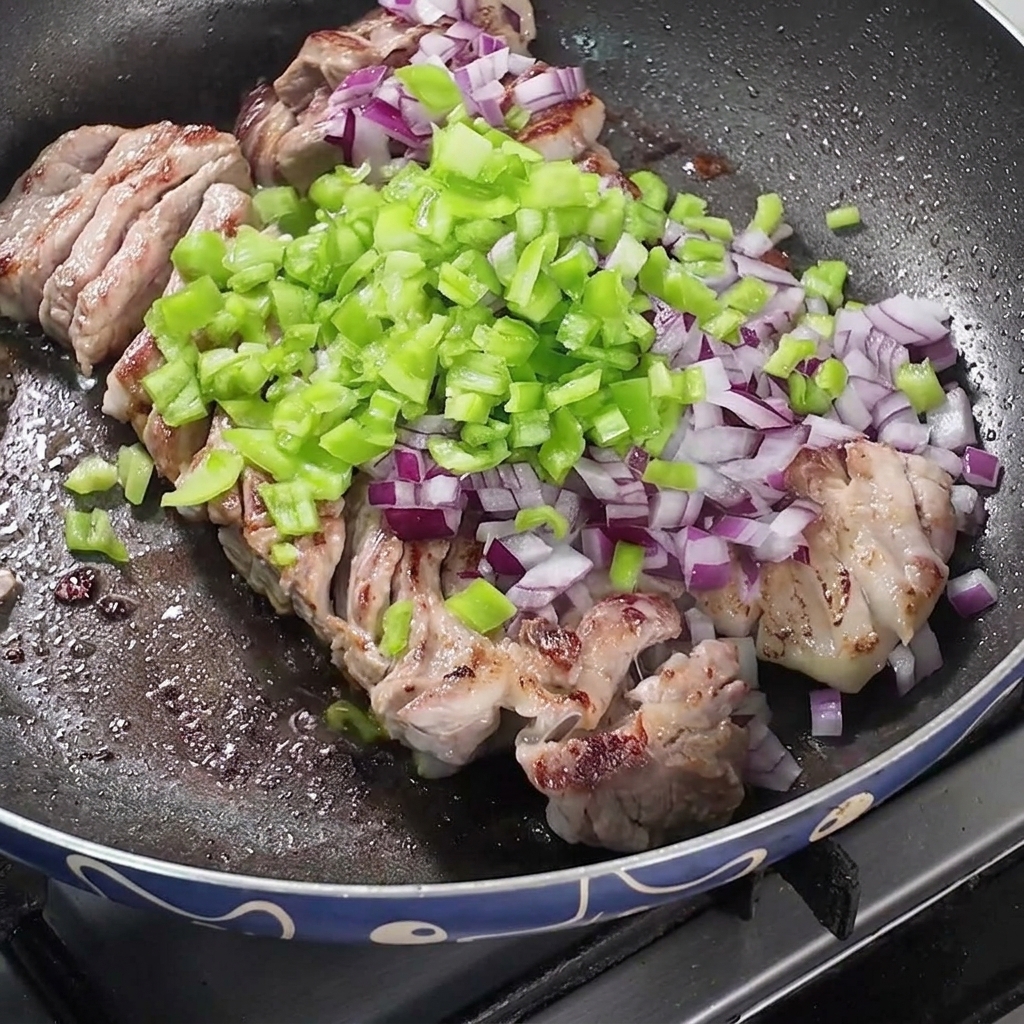 A pan filled with frying pork belly topped with piles of chopped red onions, green peppers, and dry spices.