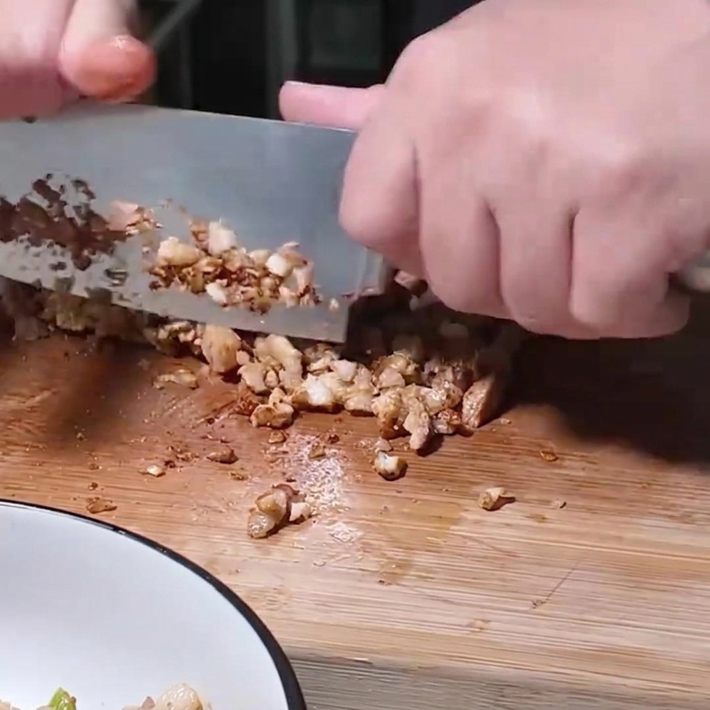 A person using a large knife to finely chop cooked pork pieces on a wooden board next to a mixing bowl.