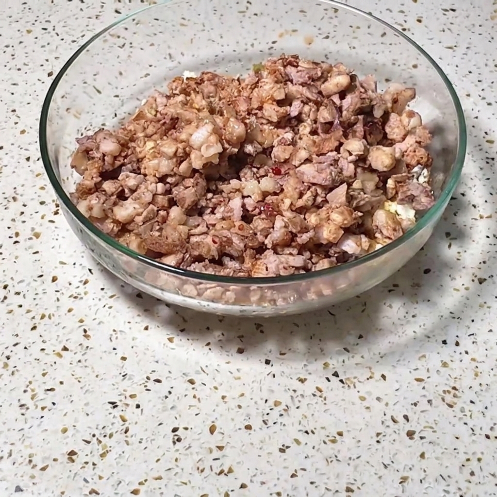 A large clear glass bowl filled with a mixture of chopped meats, eggs, and seasoned vegetables resting on a speckled countertop.