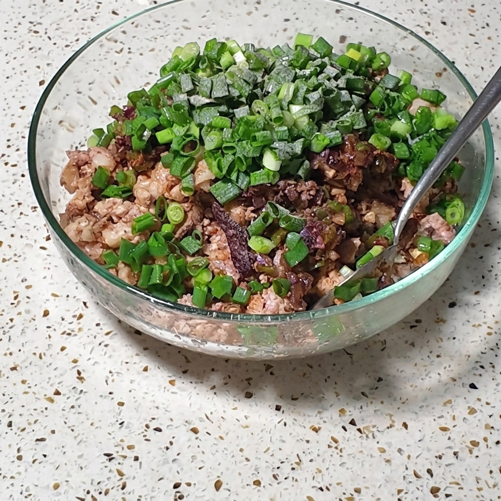A glass bowl containing a mixture of finely chopped pork and eggs, topped with fresh green onions and seasonings, being stirred with a metal spoon.