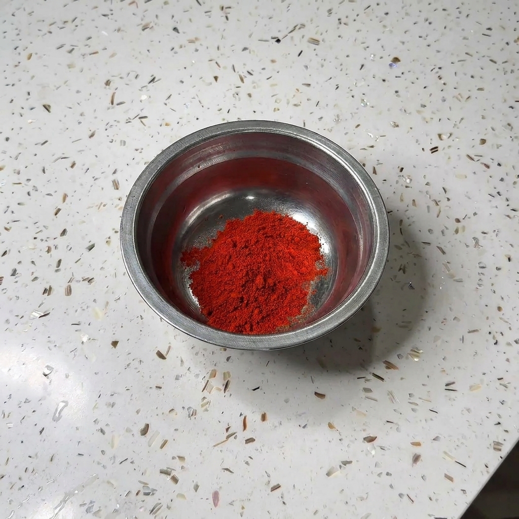A small metal bowl containing a mound of red chili powder and white sugar on a granite countertop.