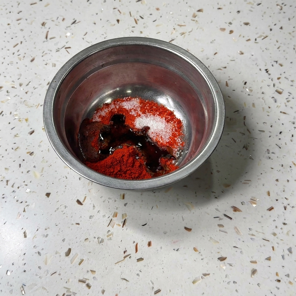 Dark liquid soy sauce and oyster sauce being poured into the metal bowl with chili powder and sugar.