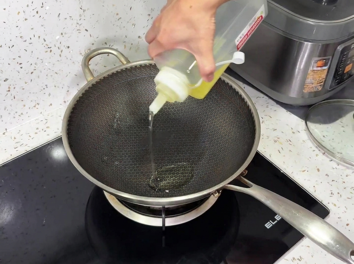 Cooking oil being poured from a squeeze bottle into a textured metal wok on a black induction cooktop.
