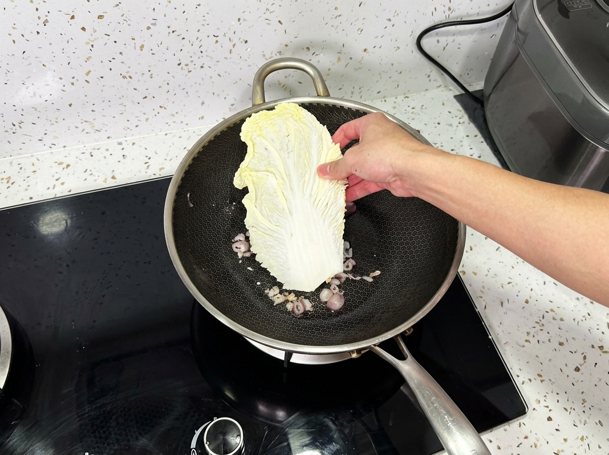 A hand placing a large leaf of napa cabbage into a wok containing cooked onions.