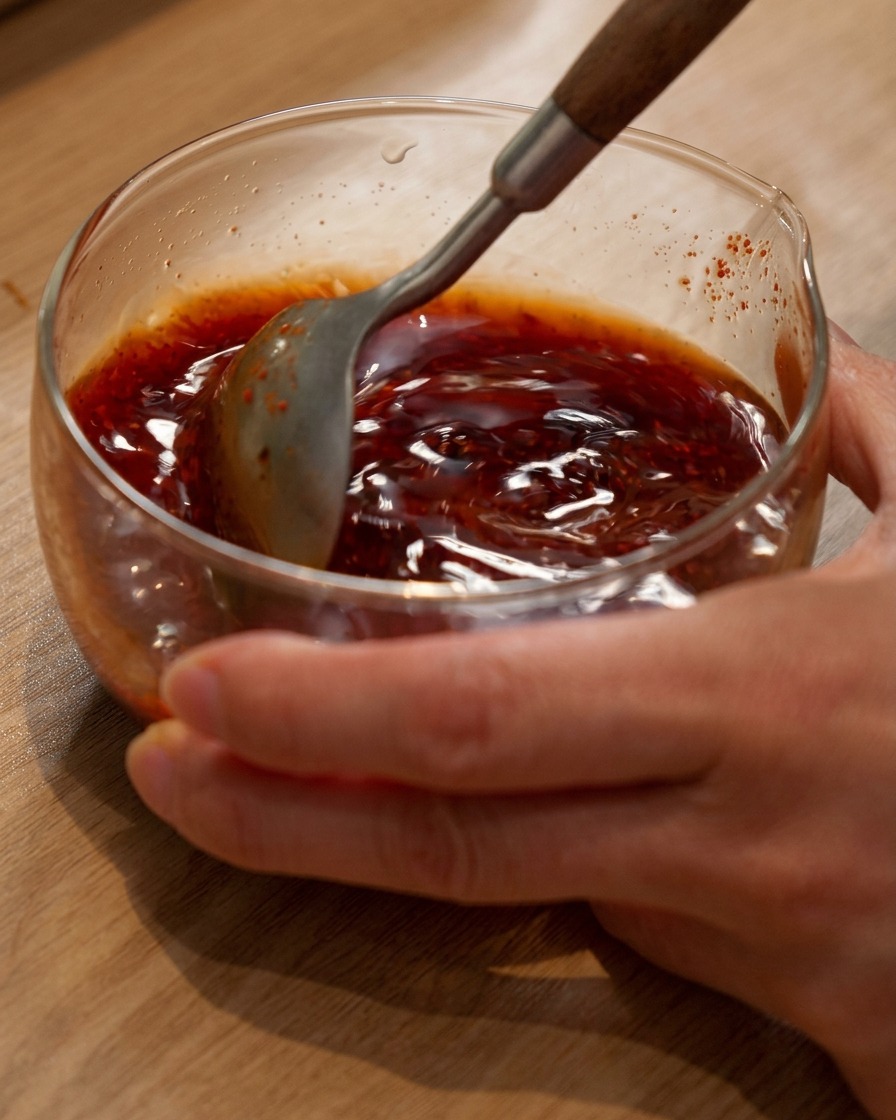 A spoon stirring a reddish-brown hot pot sauce mixture in a glass bowl.