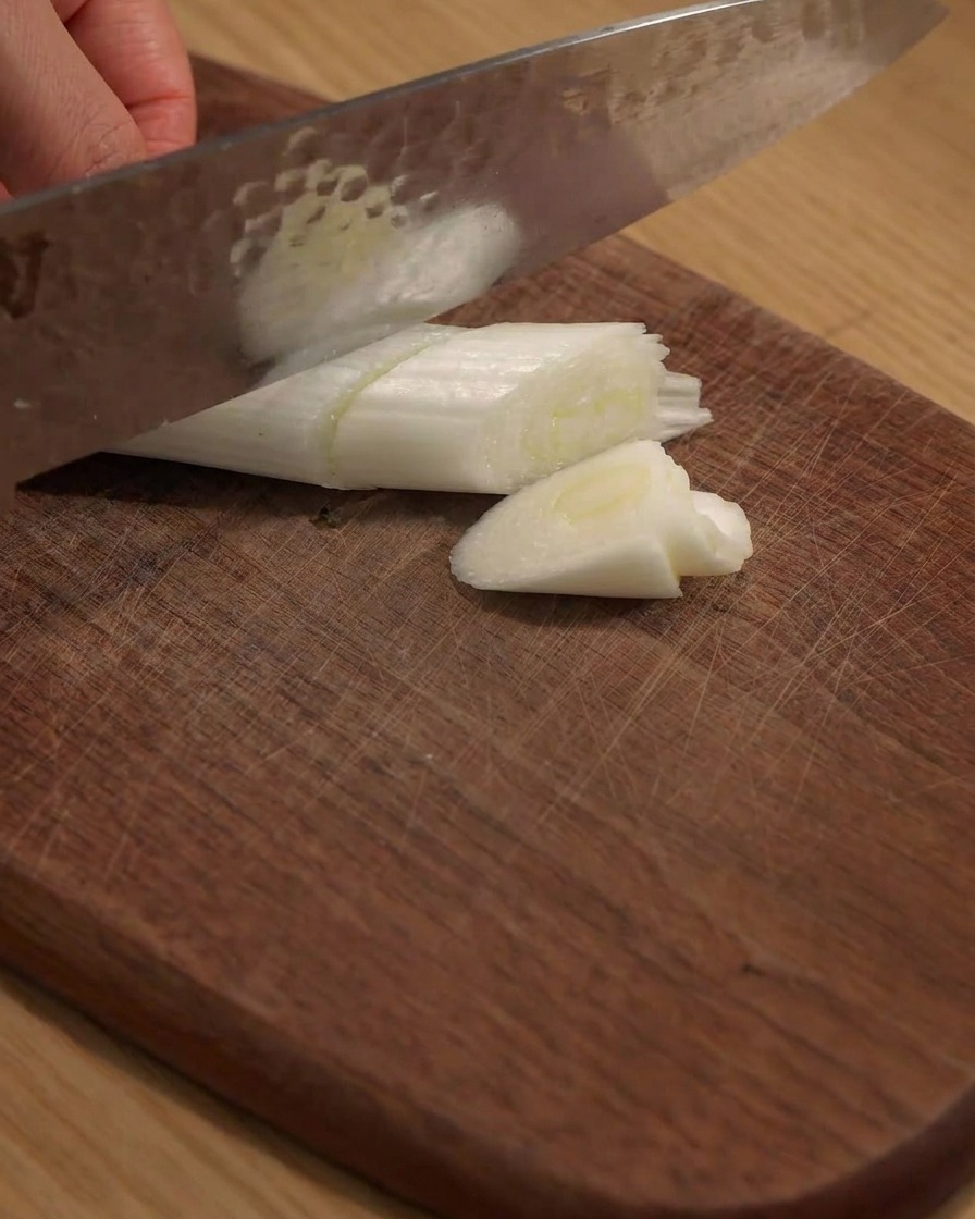 A knife cutting a large green onion into pieces on a wooden board.