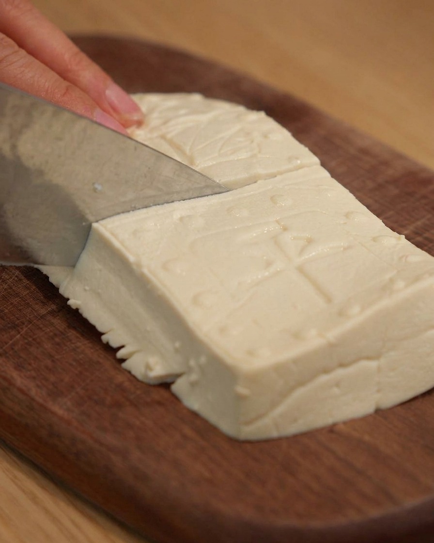 A sharp knife slicing firm tofu into neat rectangular blocks on a wooden cutting board.