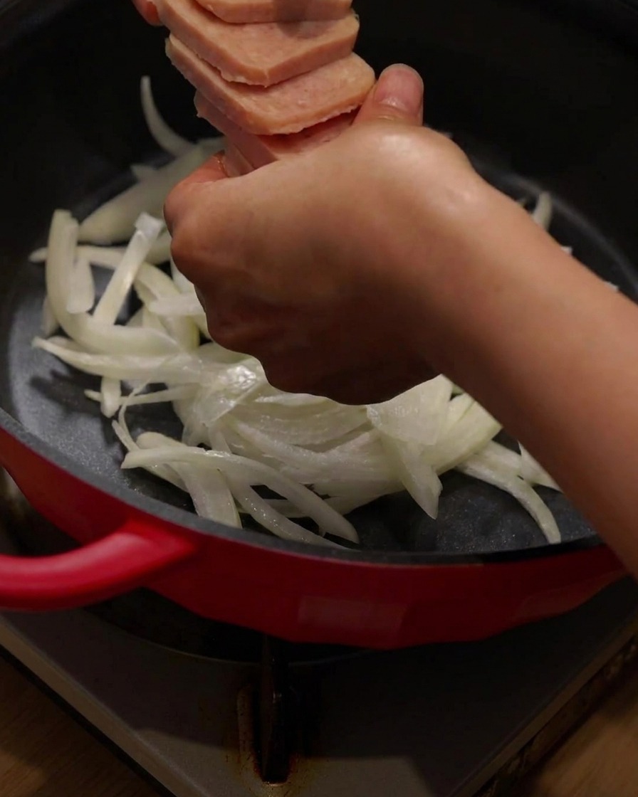 Arranging sliced luncheon meat and bacon on top of onions in the red cooking pot.