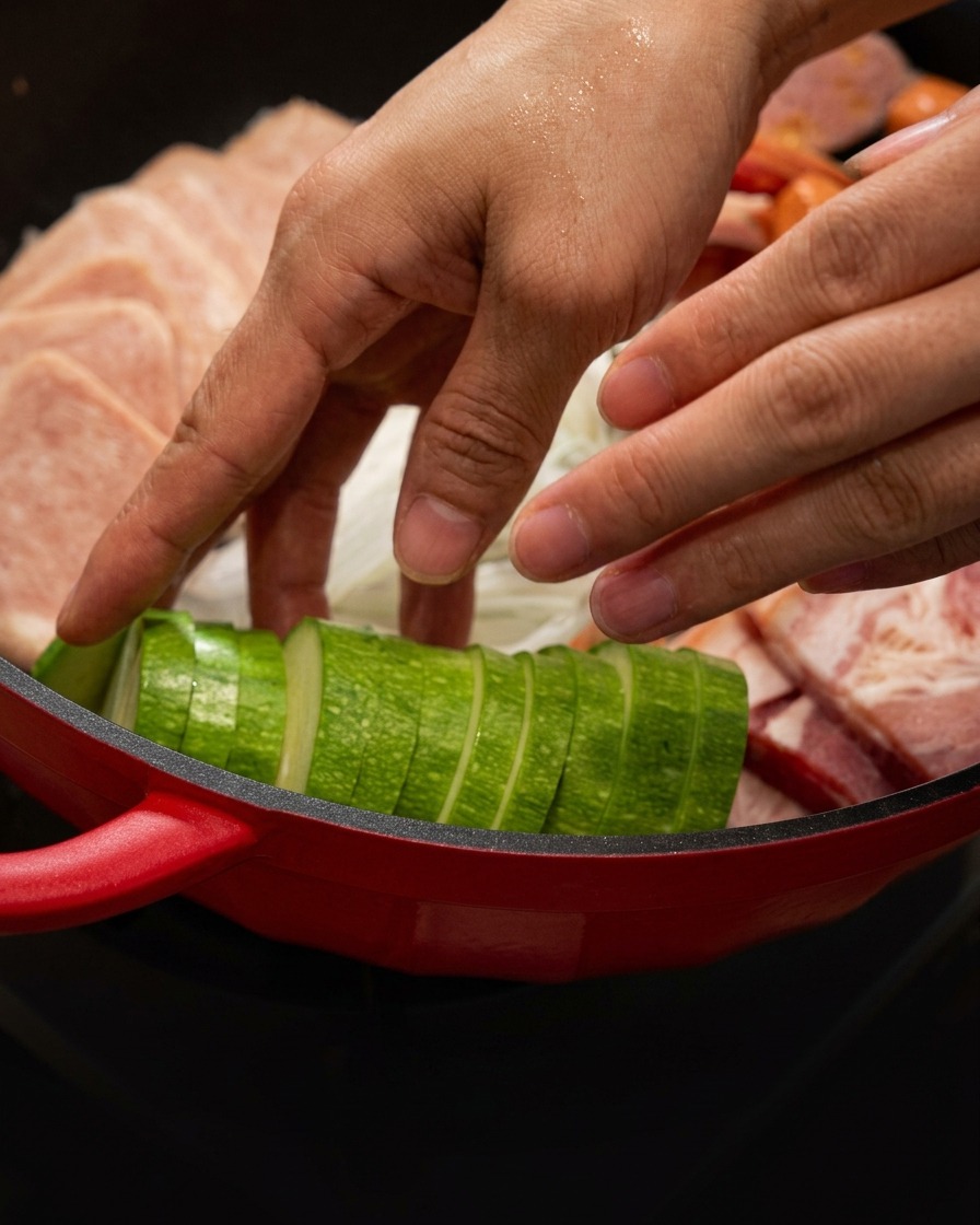 Zucchini slices being neatly placed into the red pot alongside meat.