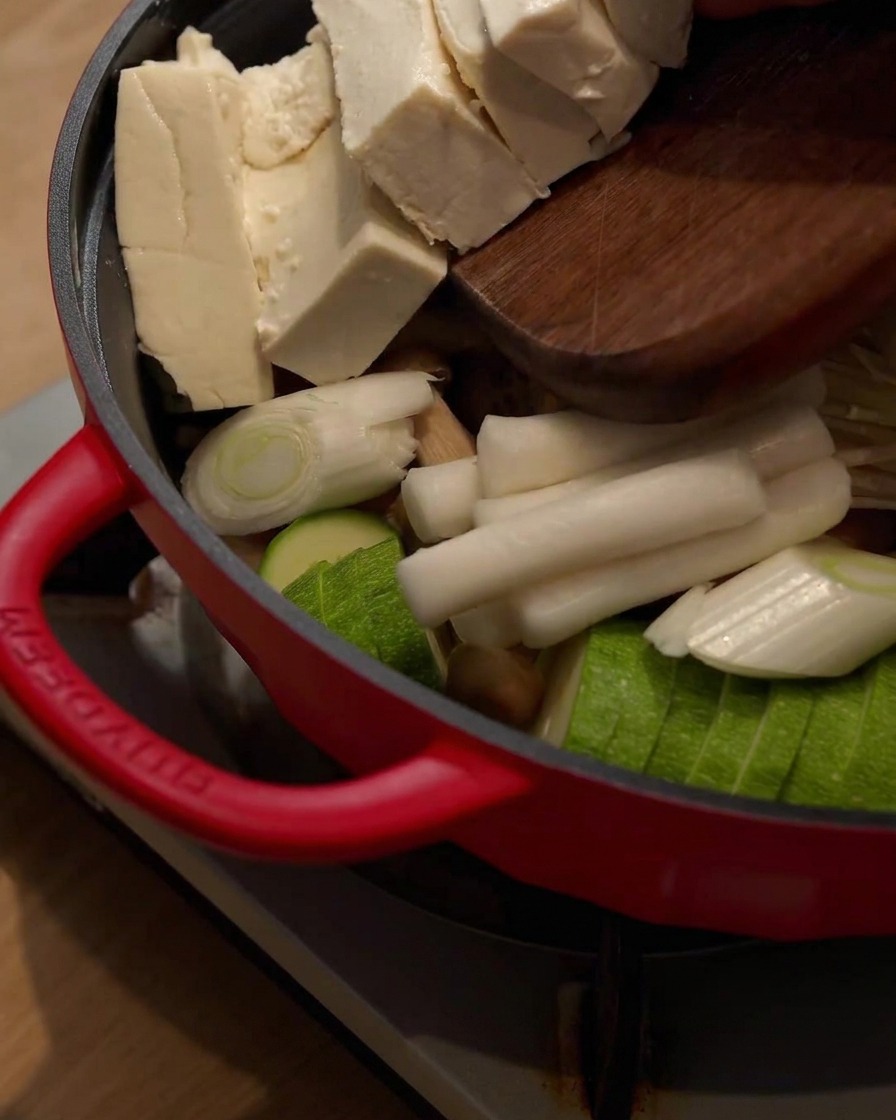 Tofu blocks being slid from a wooden cutting board into the hot pot containing other ingredients.