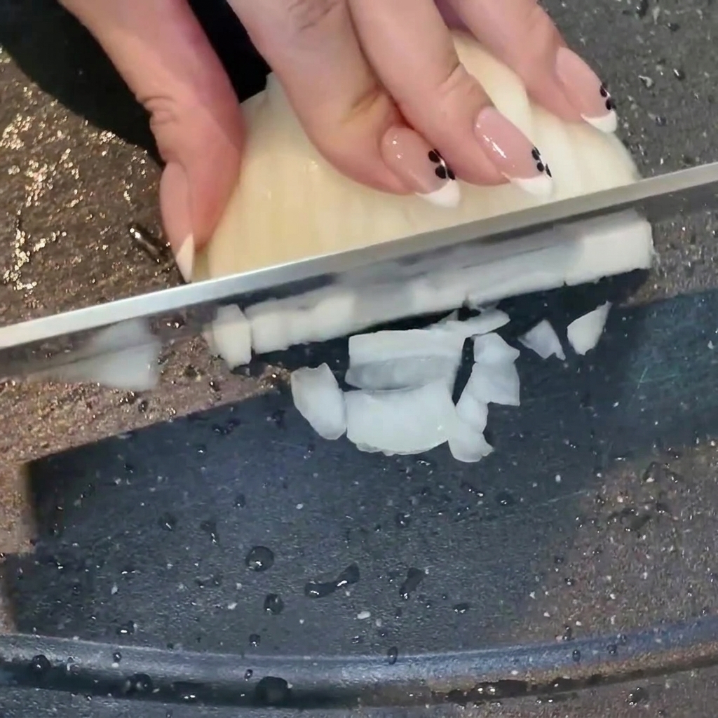 A hand using a knife to dice a white onion on a dark cutting board, with minced garlic nearby.