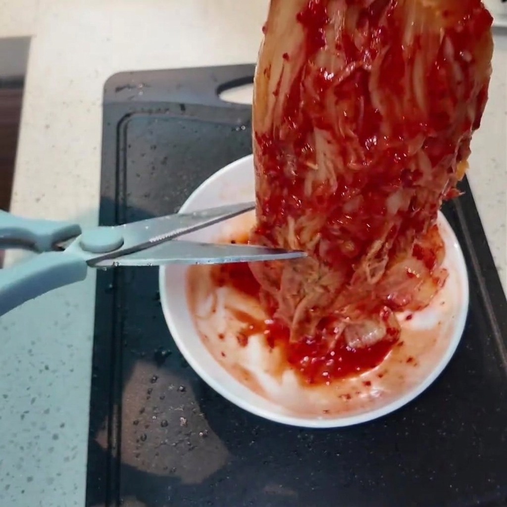 A piece of vibrant red kimchi being snipped into smaller pieces with kitchen scissors over a small white dish.